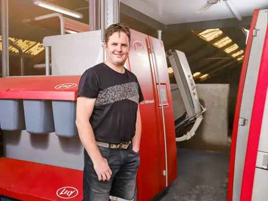 A person stands smiling in a modern dairy farm, wearing a black t-shirt and denim shorts. Behind them, red automated milking machines are visible, with bright lighting and a clean, organized environment.